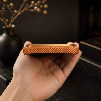 A hand holds an orange woven case nearly edge-on, showing the fine textured rim and bottom charging and speaker openings against a warm dark background.