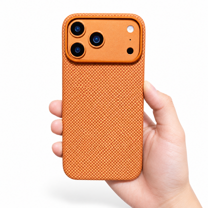 A hand holds a bright orange fine-weave phone case upright on white. A broad matching camera panel with three black lenses sits above the densely interlaced back pattern. A thin gold-orange rim outlines the rounded corners and camera panel, and the hand supports the lower edge.