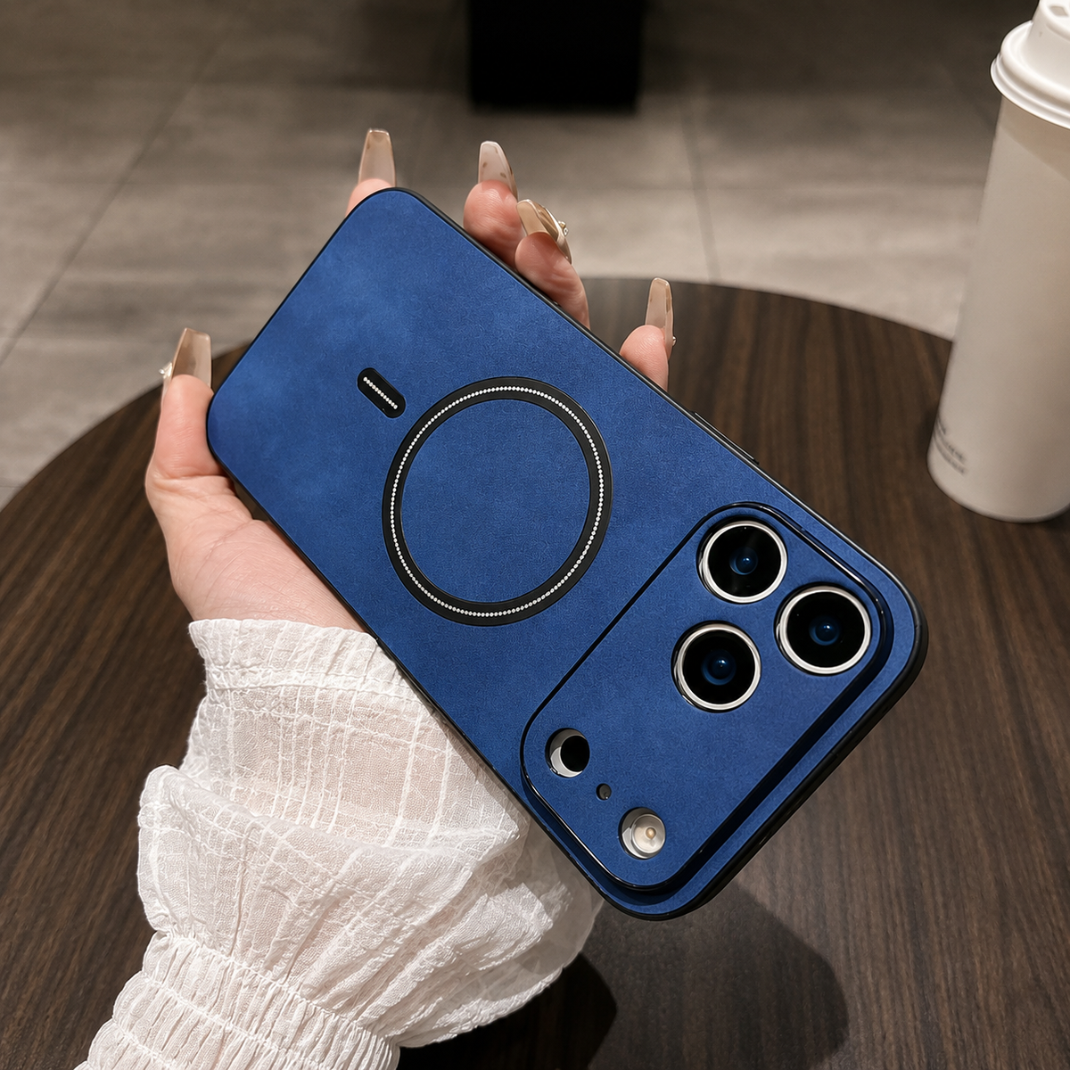 Blue leather-look phone case held diagonally in a manicured hand above a wooden table, with a black ring and matching wide camera guard.