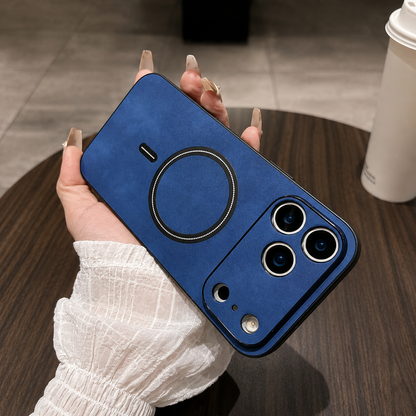 Blue leather-look phone case held diagonally in a manicured hand above a wooden table, with a black ring and matching wide camera guard.