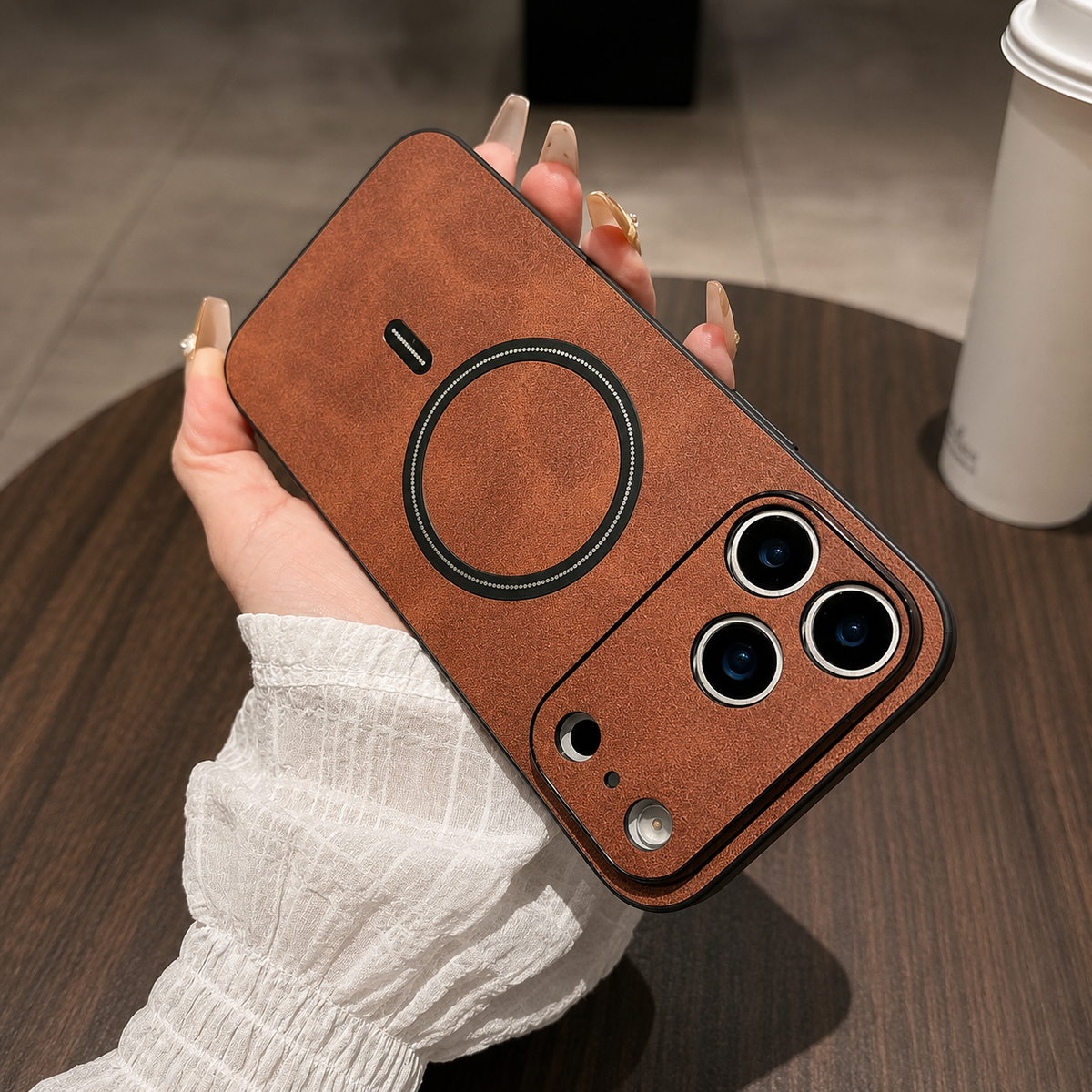 Tan leather-look phone case held diagonally in a manicured hand above a wooden table, showing its black ring and raised camera guard.