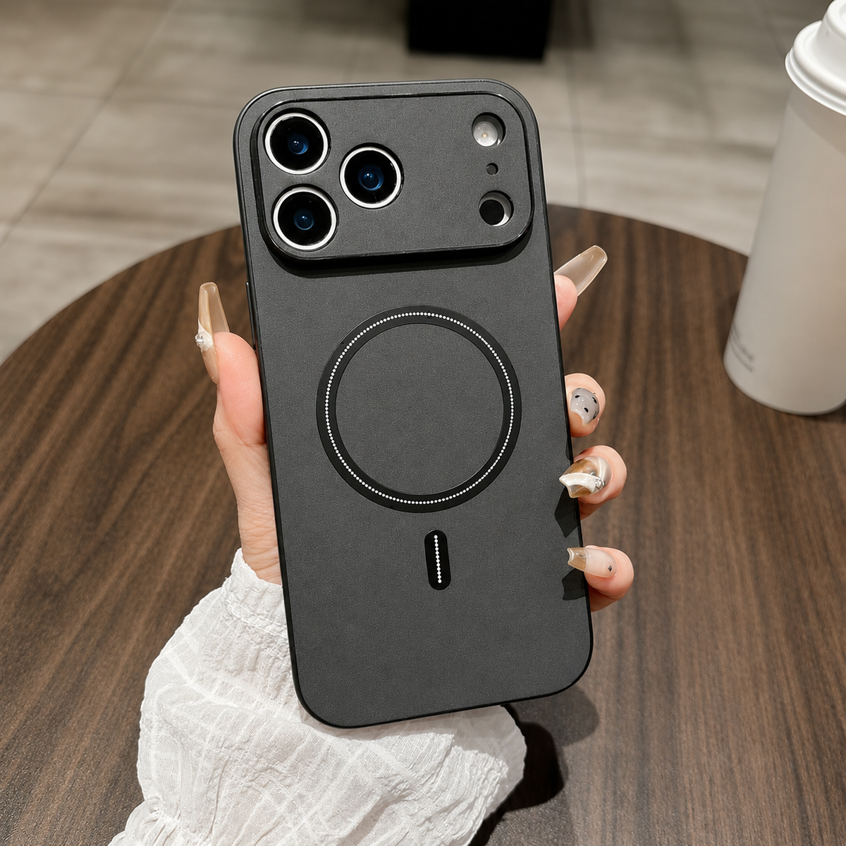 A hand holds a charcoal leather-look phone case above a round wooden table, with a raised camera panel and black circular back ring.