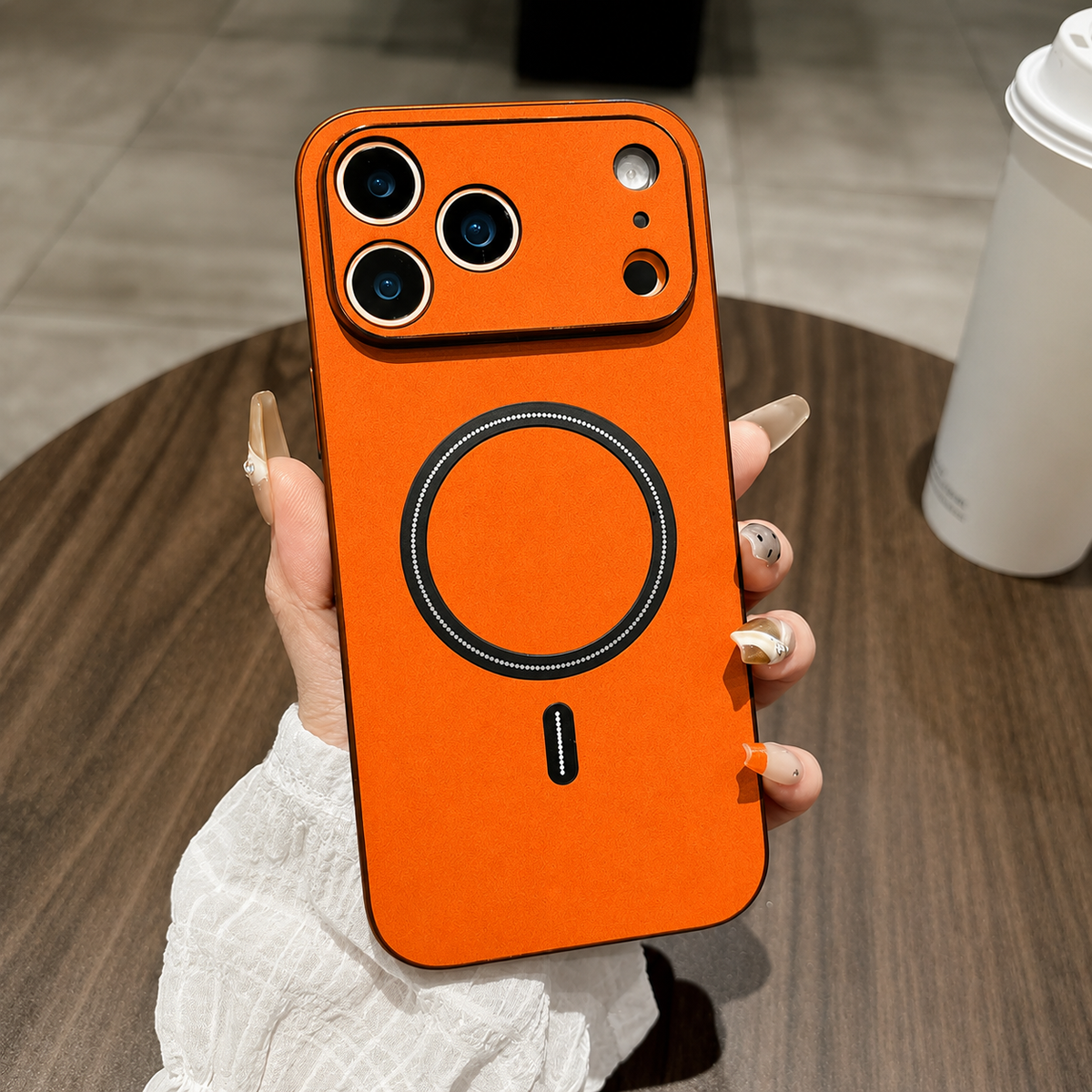 A hand holds a bright orange leather-look phone case above a wooden table, with black edges, a matching camera guard and black ring and bar.