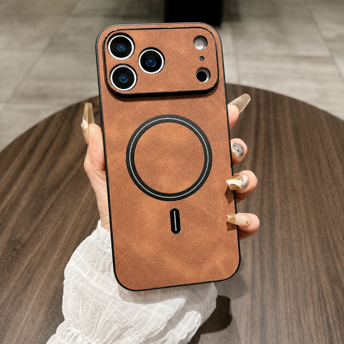 A hand holds a tan leather-look phone case above a wooden table, with a matching wide camera guard and black circular back ring.