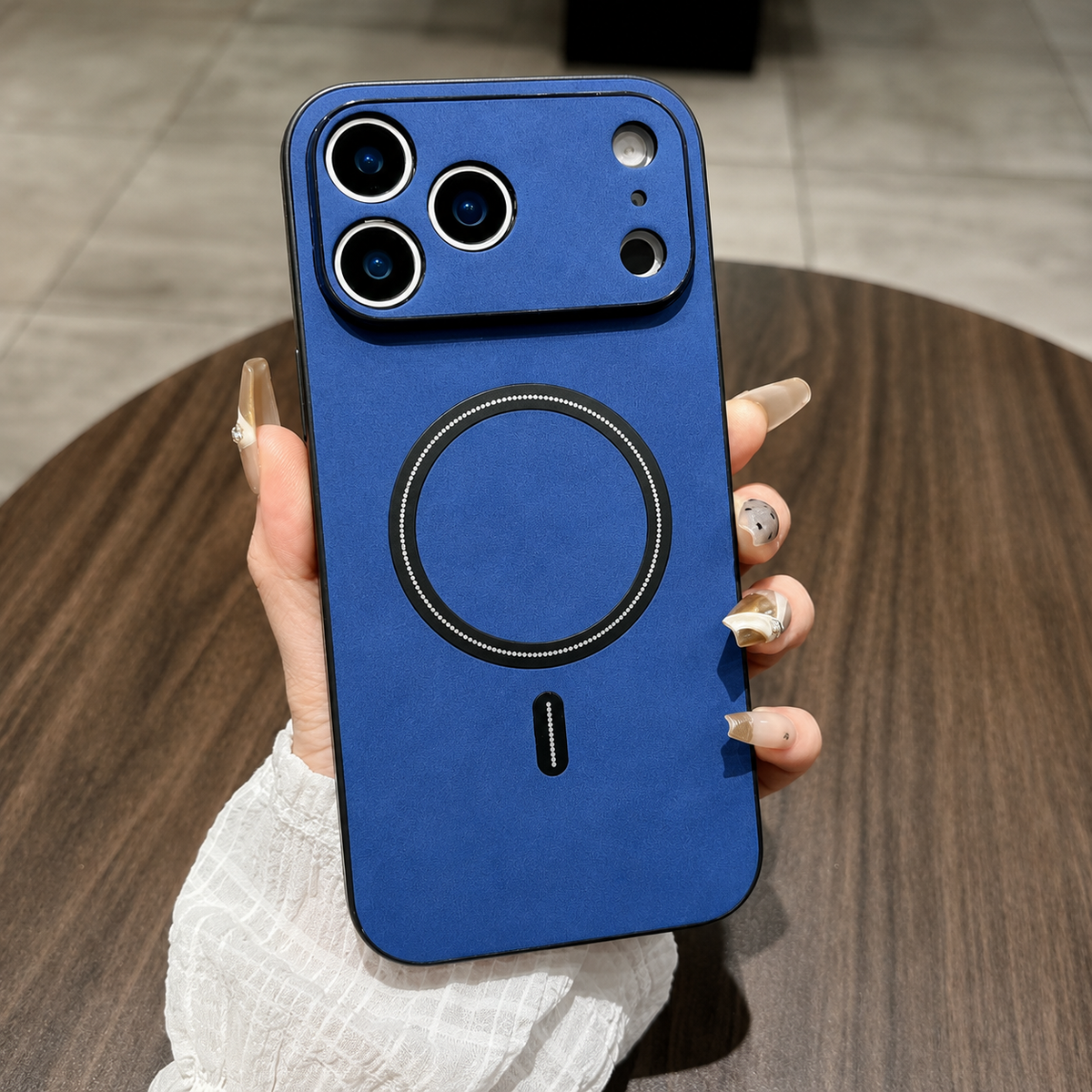 A hand holds a blue leather-look phone case above a wooden table, with black edges, a matching camera panel and black circular ring and bar.