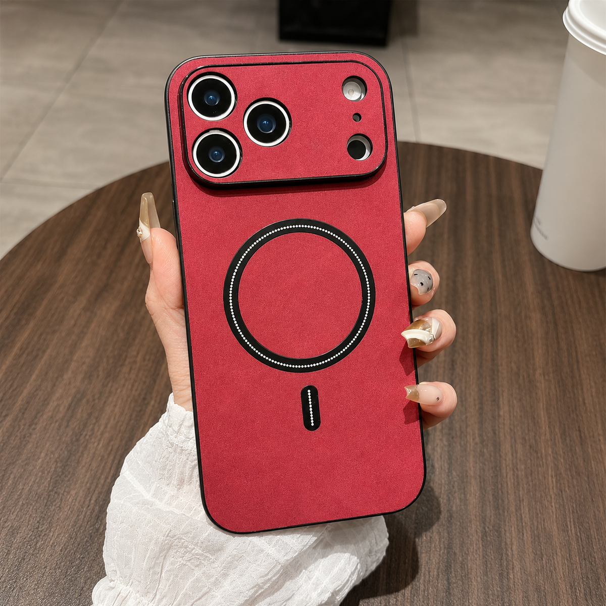A hand holds a red leather-look phone case above a wooden table, with a matching camera guard, black border and black circular ring and bar.