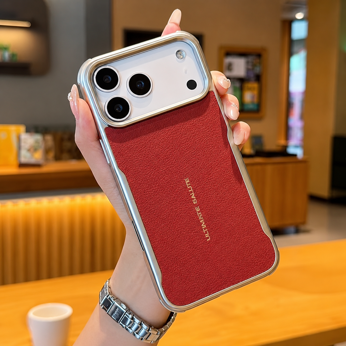 A hand holds a red leather-look phone case with reflective metallic edging, a wide camera opening and small gold lettering in a café.