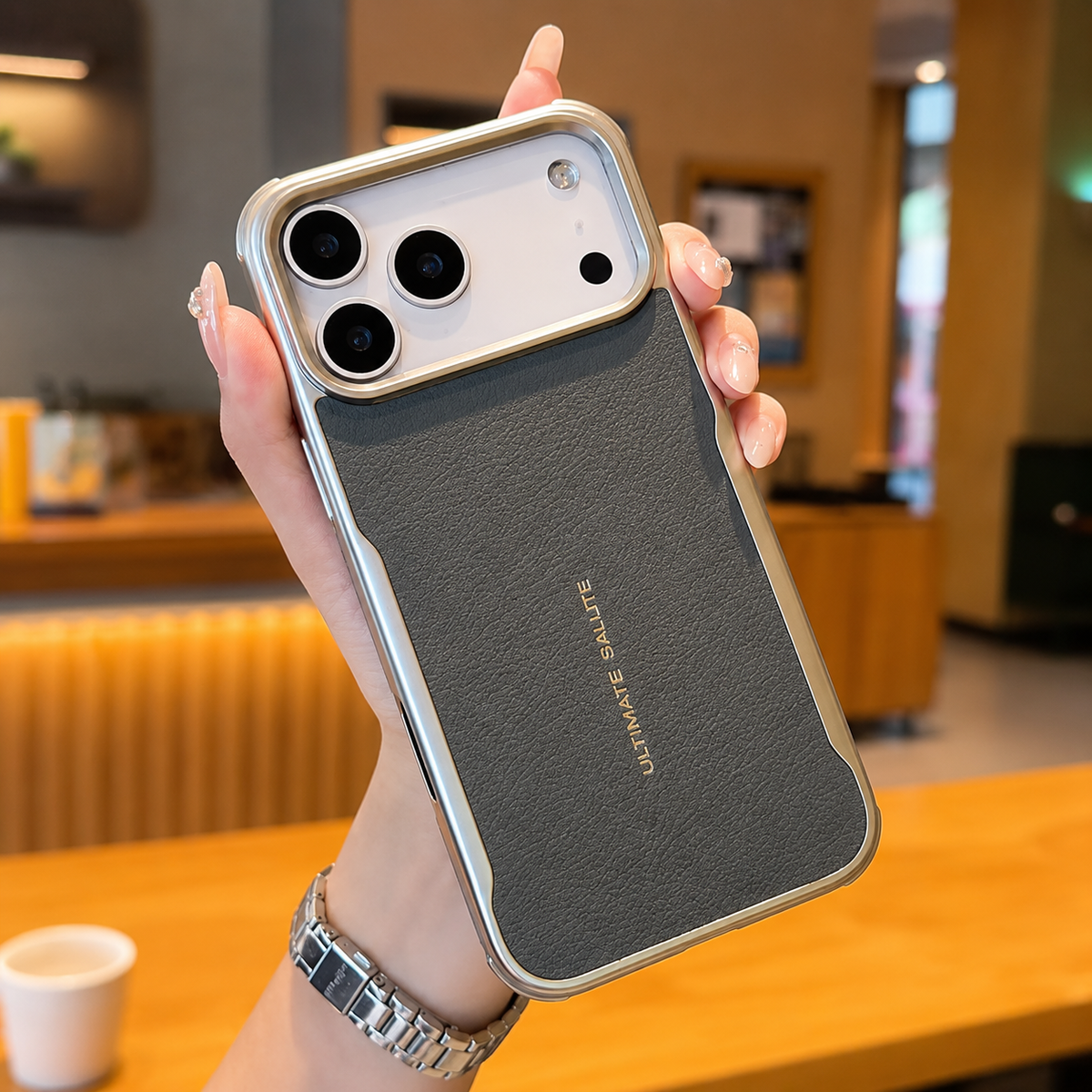 A hand holds a gray leather-look phone case with reflective edging and gold 'Ultimate Salute' lettering in a café.