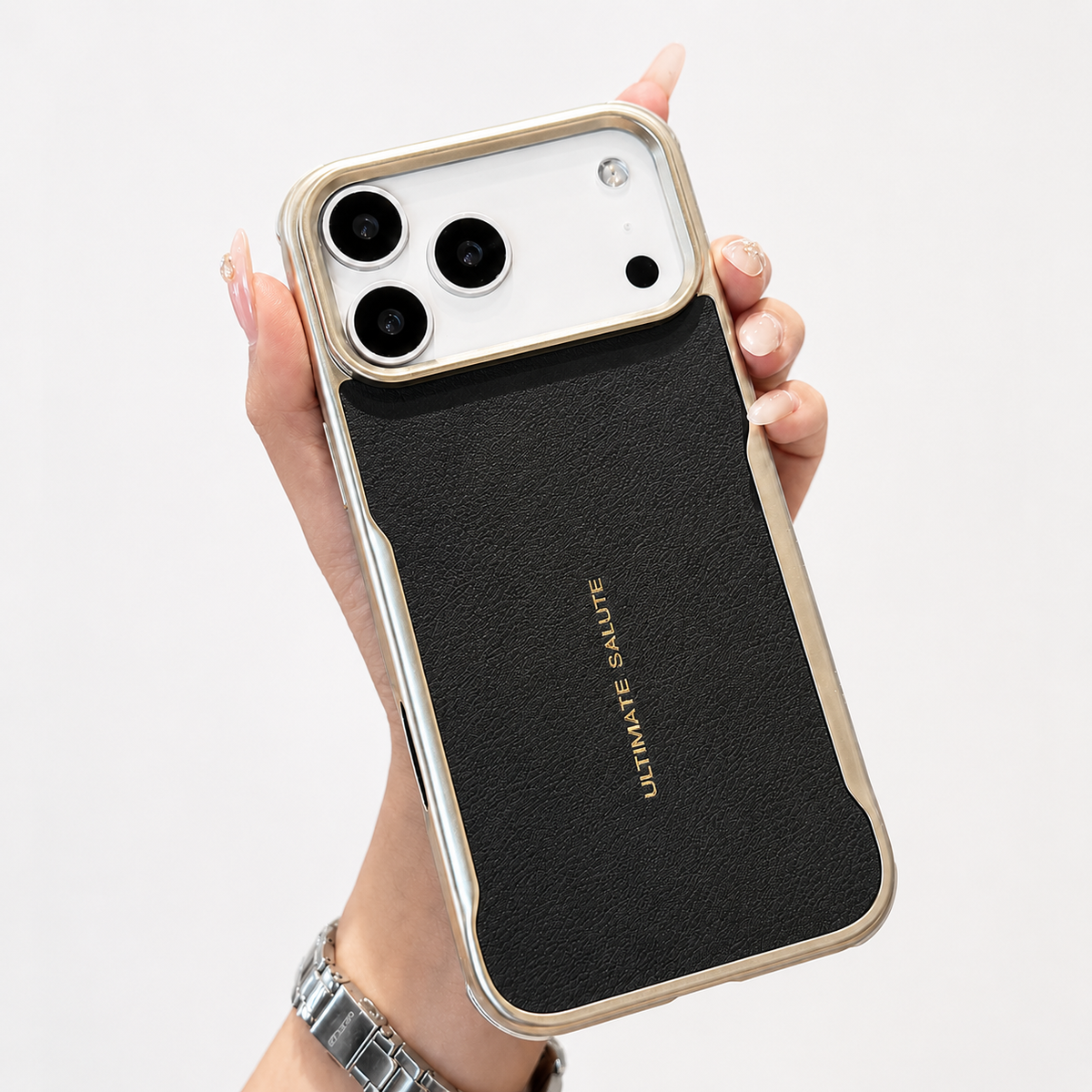 A hand wearing a silver watch holds a black leather-look phone case with reflective metallic edging, a wide camera opening and small gold 'Ultimate Salute' lettering.