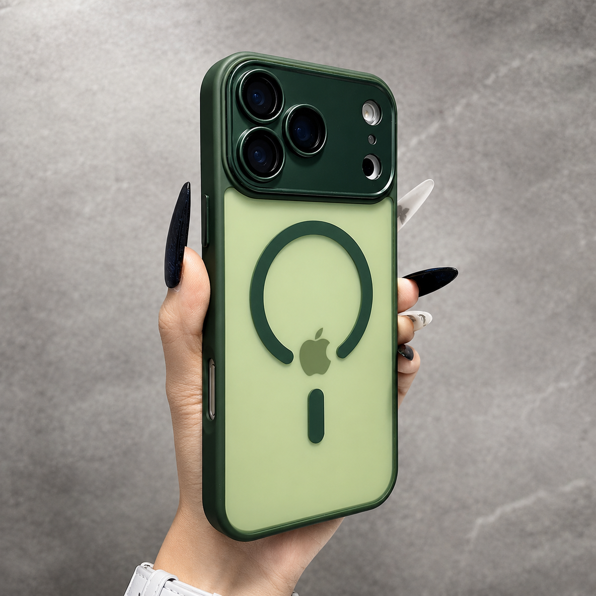 A hand holds a pale green frosted phone case with dark green trim against gray stone. Its dark green ring surrounds the faint Apple logo beneath the camera panel.