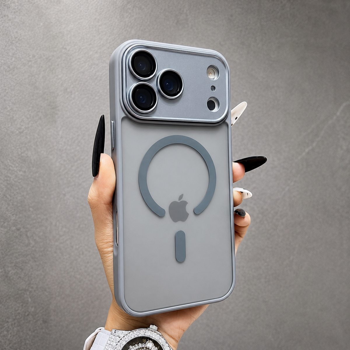 A hand wearing a jeweled watch holds a gray frosted phone case against gray stone, displaying a matching rear ring, short bar and broad camera panel.