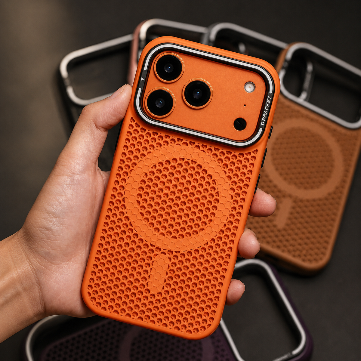 A hand displays an orange perforated phone case upright, showing its circular rear ring and folded camera-frame stand against a background of other cases.