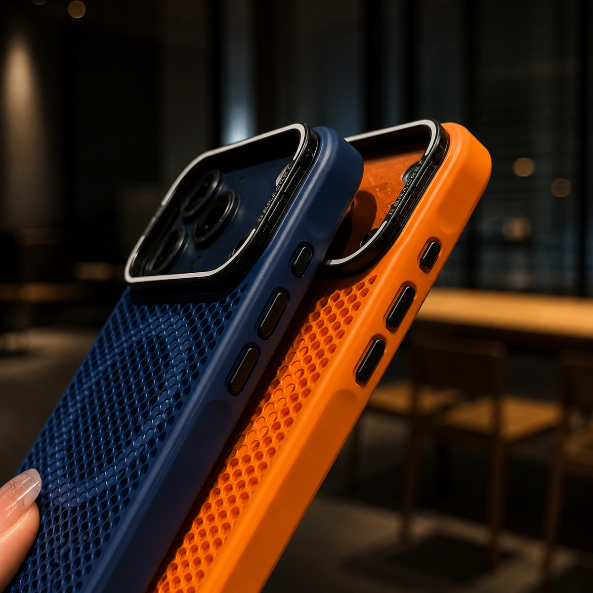 Navy and orange perforated phone cases are held edge-on, revealing black side buttons and raised silver-edged camera frames in a dim café setting.