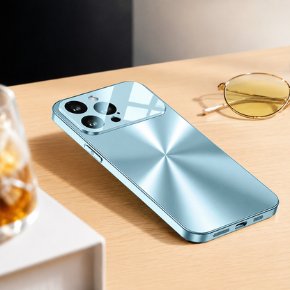 A pale blue metallic-looking phone case lies on a wooden table beside yellow-tinted glasses and a drink. Its back has a radial brushed pattern and reflective camera panel.