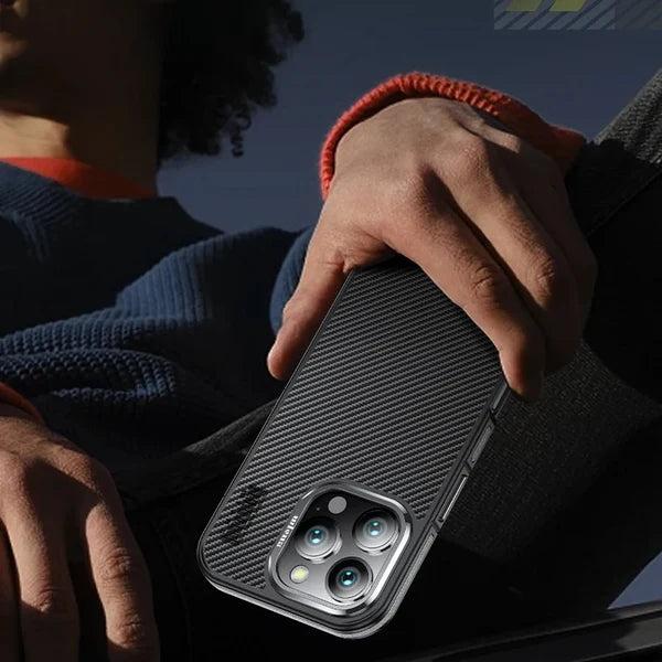 A cropped person in dark blue knitwear and a black jacket holds a black woven phone case diagonally against their torso outdoors. The back faces the viewer, showing a fine diagonal pattern and a pale three-lens camera surround near the lower end of the image. An orange bracelet circles the wrist, with blue sky behind.