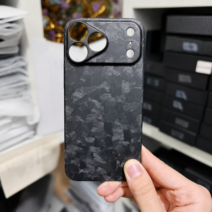 A hand holds a matte black shard-patterned case in front of stacked boxes. Its broad camera panel has separate circular lens and sensor openings.
