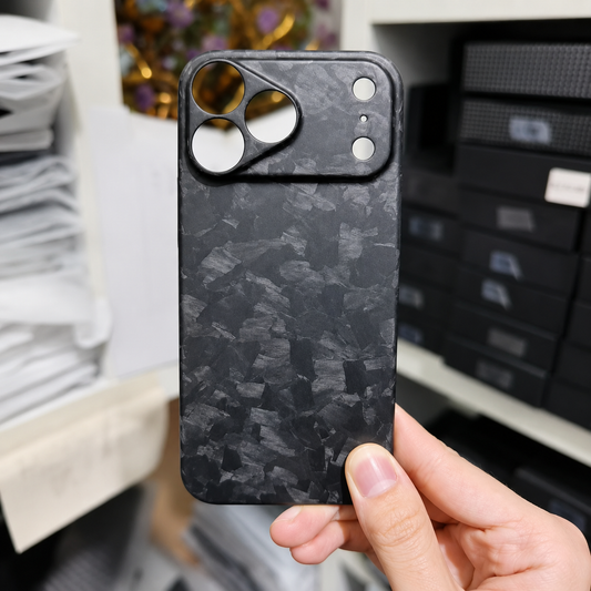 A hand holds a matte black shard-patterned case in front of stacked boxes. Its broad camera panel has separate circular lens and sensor openings.