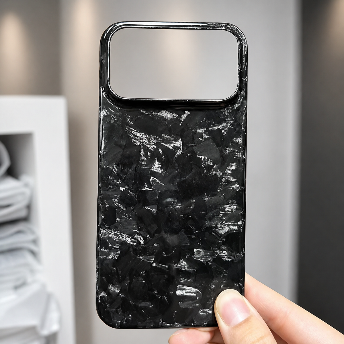 A hand holds a glossy black case with silver-gray shard-like patches in front of shelves. A wide rectangular camera opening spans the top.