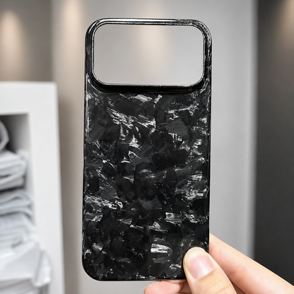 A hand holds a glossy black case with silver-gray shard-like patches in front of shelves. A wide rectangular camera opening spans the top.