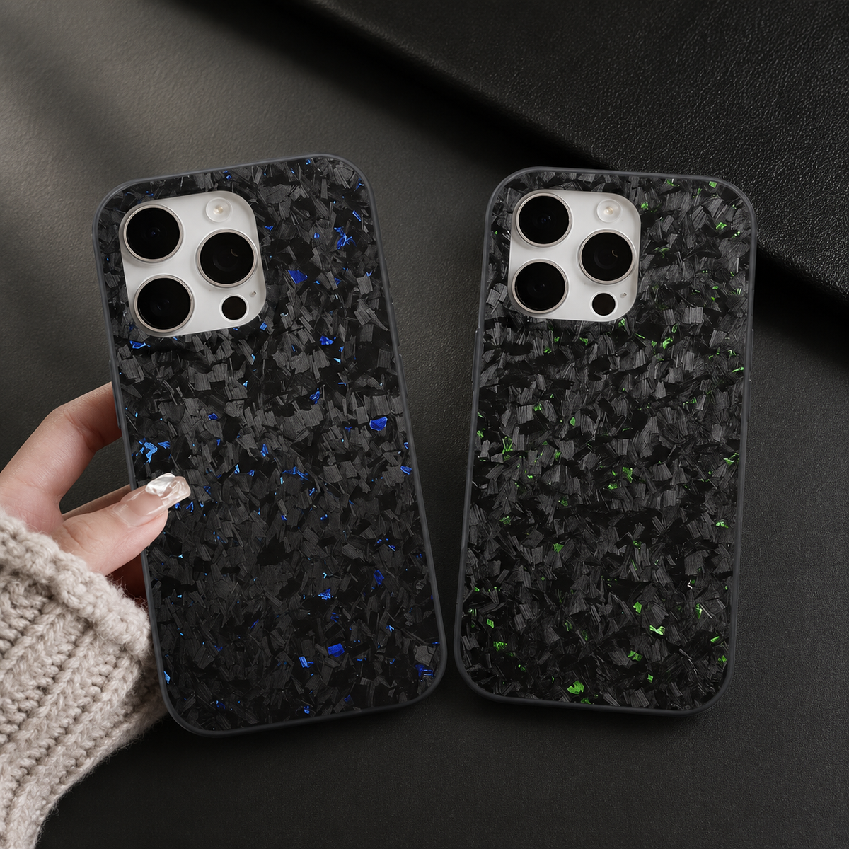 A hand in a cream knit sleeve holds blue- and green-flecked black cases side by side above a dark surface. Pale camera panels contrast with the irregular dark mottled backs.