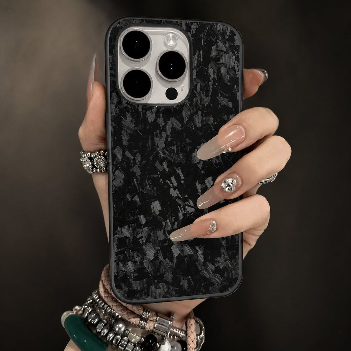 A hand with long pale decorated nails and stacked bracelets holds a gray-flecked black case upright. Its white camera panel contains three large dark lenses. The background is softly blurred.