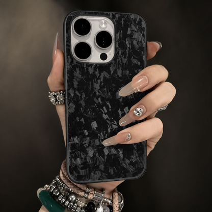 A hand with long pale decorated nails and stacked bracelets holds a gray-flecked black case upright. Its white camera panel contains three large dark lenses. The background is softly blurred.