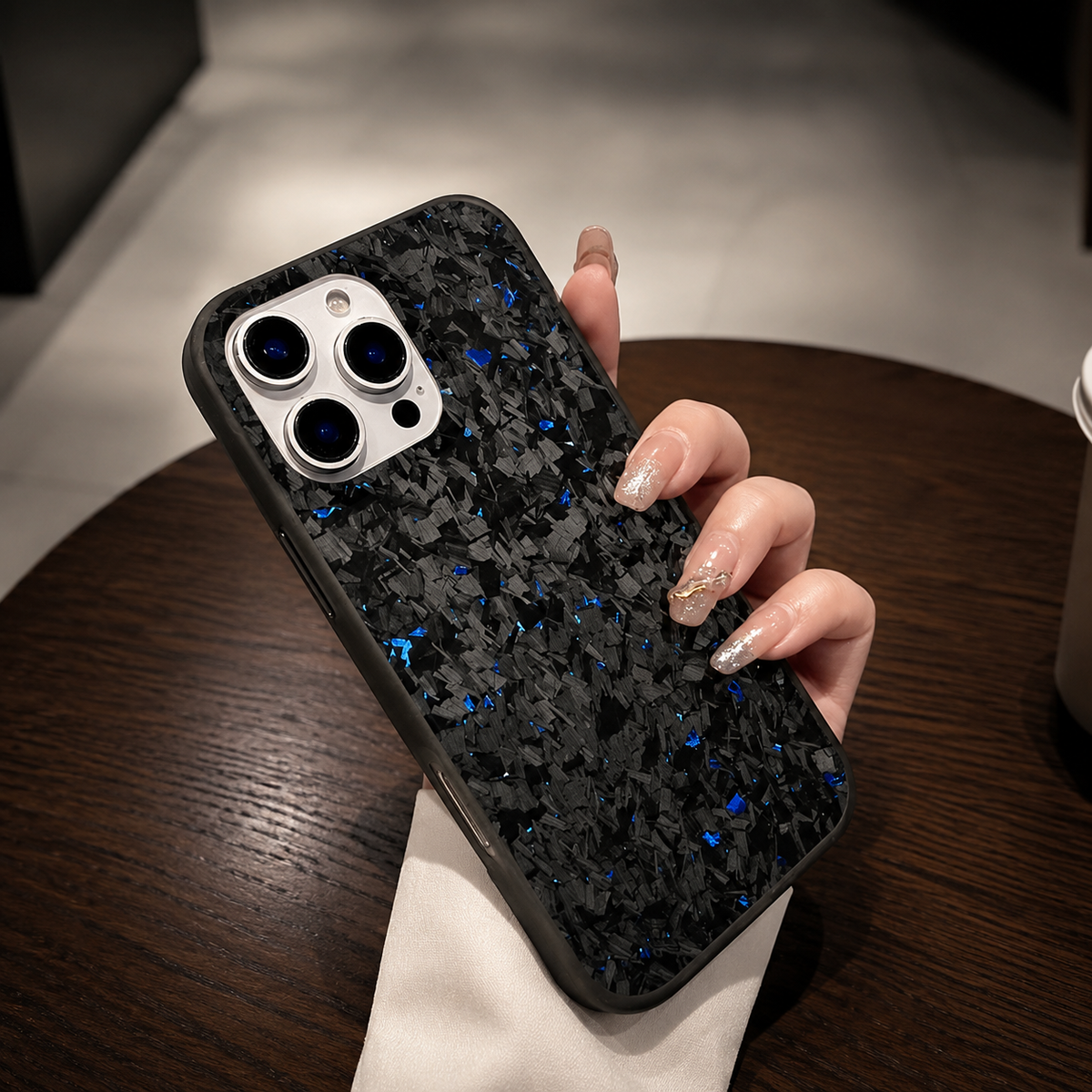 A hand with pale nails holds a blue-flecked black case diagonally above a round wooden café table. The pale camera panel and mottled back face forward; a cream sleeve frames the wrist.