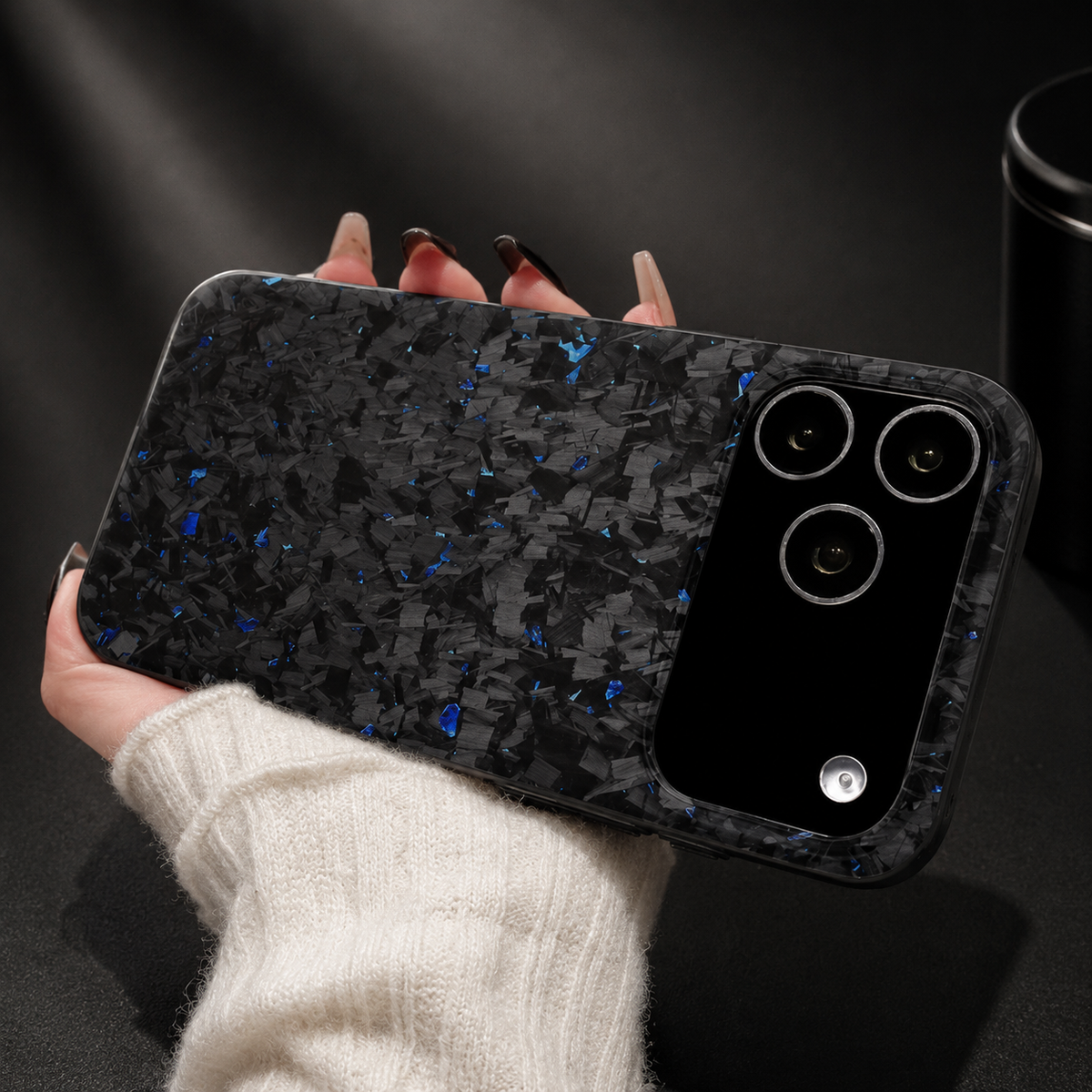 A hand in a cream knit sleeve holds a blue-flecked black case horizontally against dark fabric. Its broad black camera panel sits at the right, while irregular blue flecks scatter across the back.