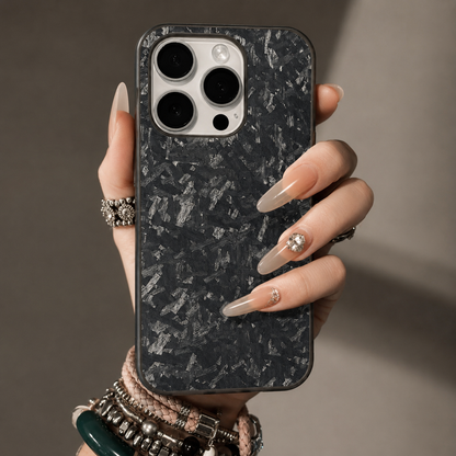 A hand with long pale decorated nails and stacked bracelets holds a gray-flecked black case upright. A pale camera panel contrasts with the mottled back. The softly lit background is gray-brown.