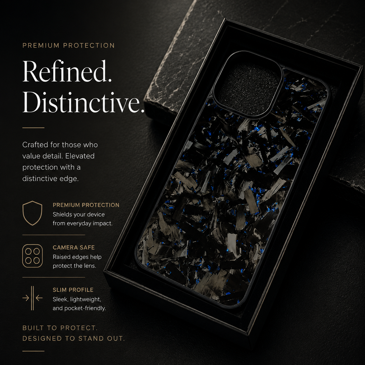 A black case with gray fragments and blue flecks lies diagonally in a black tray on stone. Its rounded square camera opening has a raised black border.