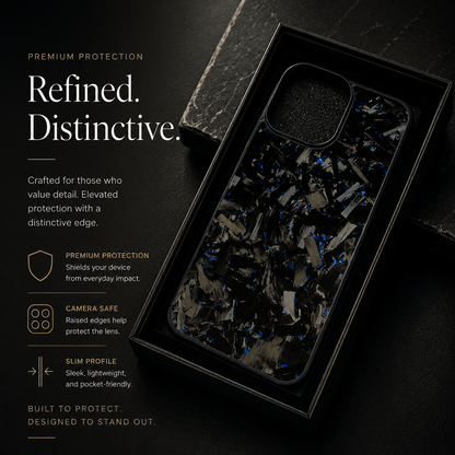 A black case with gray fragments and blue flecks lies diagonally in a black tray on stone. Its rounded square camera opening has a raised black border.