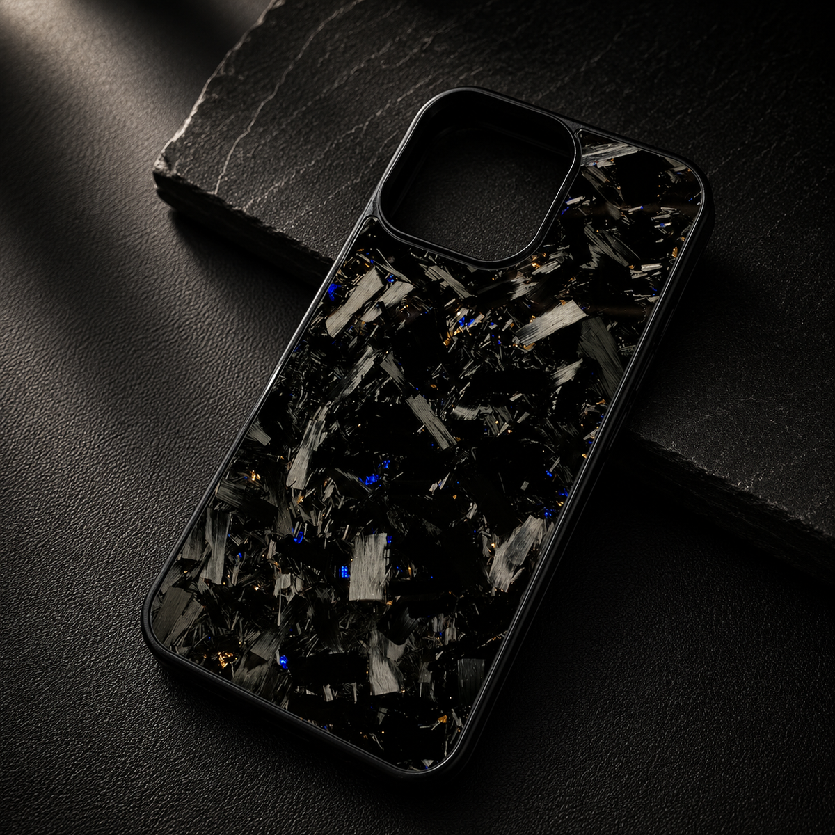 A glossy black case with large silver-gray fragments and small yellow-gold flecks rests diagonally across a dark stone slab. The raised empty camera opening remains visible.