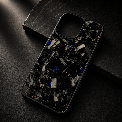 A glossy black case with large silver-gray fragments and small yellow-gold flecks rests diagonally across a dark stone slab. The raised empty camera opening remains visible.