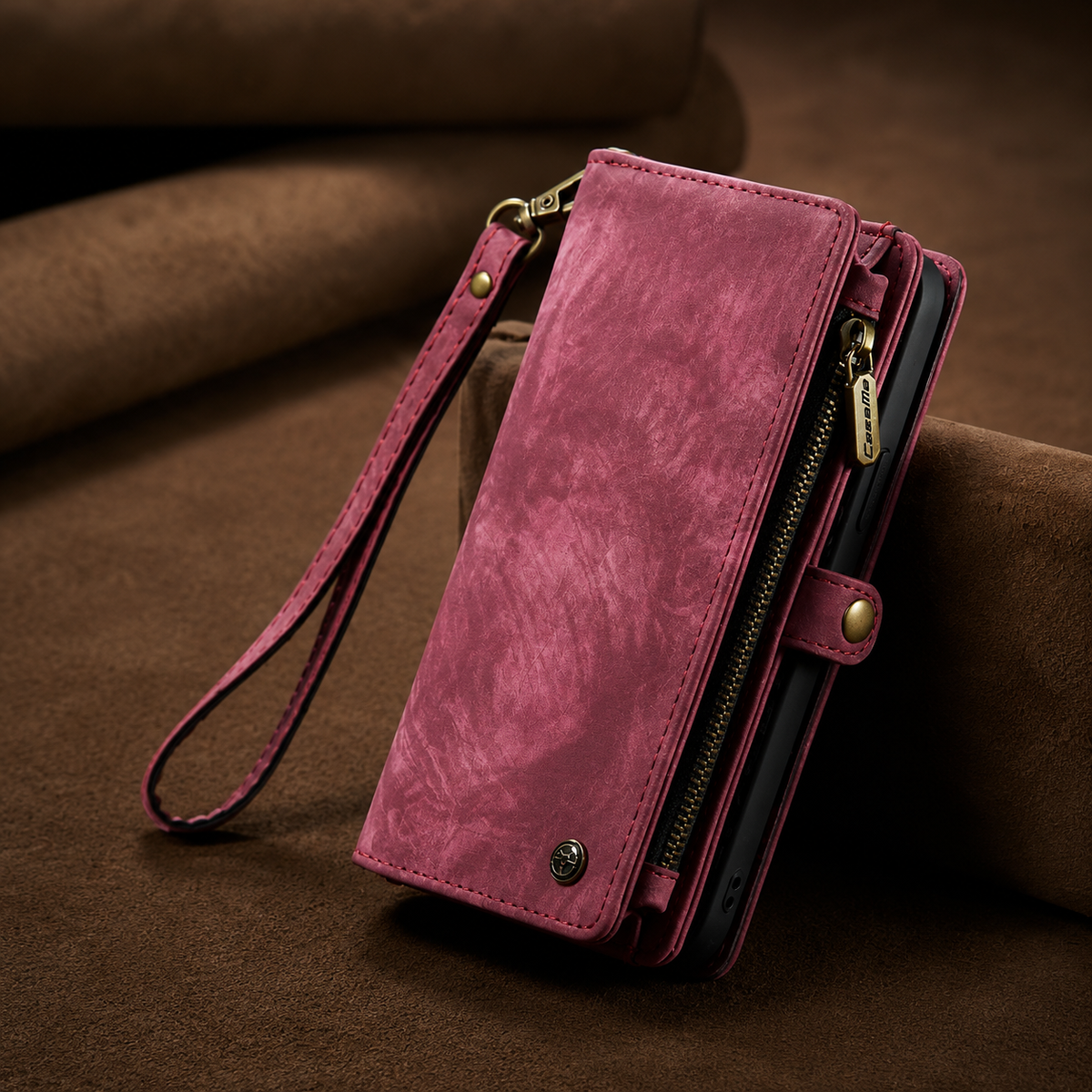 A closed burgundy leather-look wallet phone case stands on brown fabric. A wrist strap hangs at the left, and a brass-colored zipper and snap tab line the right edge.