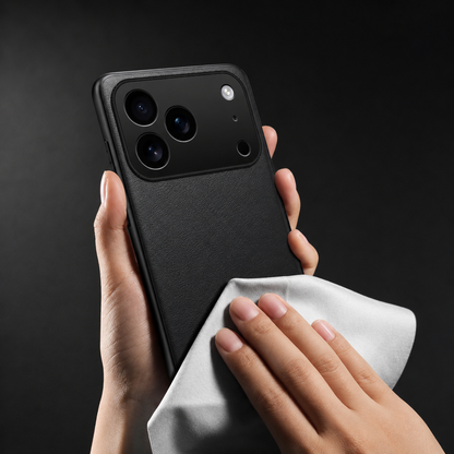 One hand holds a black phone case upright against a dark background while another wipes its fine-textured back with a folded white cloth. The broad raised camera panel contains three large lenses and two smaller circular openings.