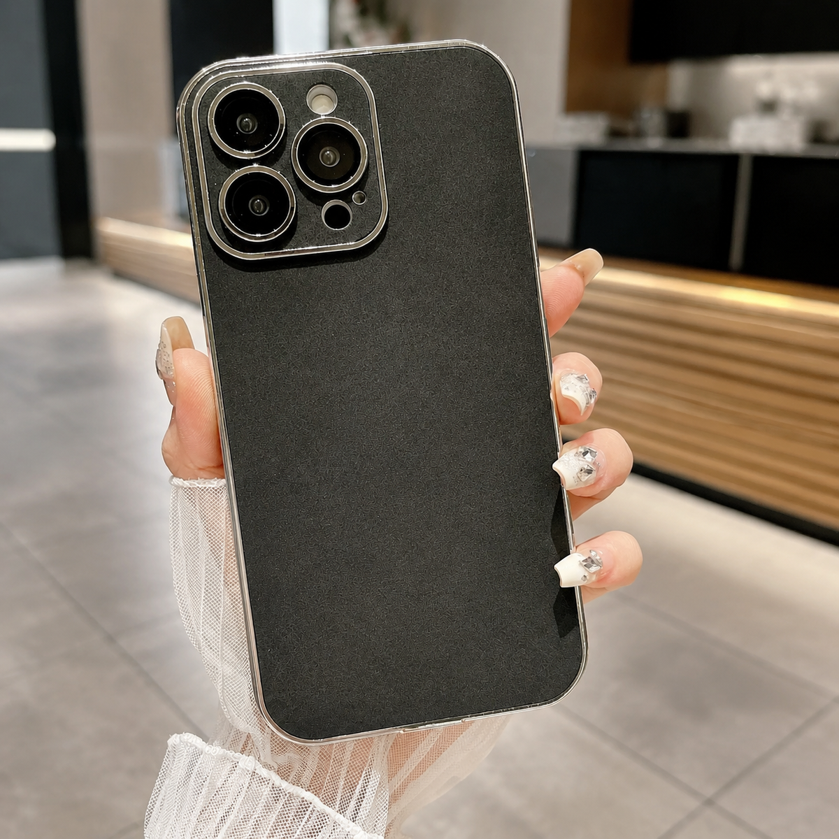 Hand holds a black suede-look case upright in a café, showing a finely mottled back, silver-colored border, and matching camera panel with individual lens openings.