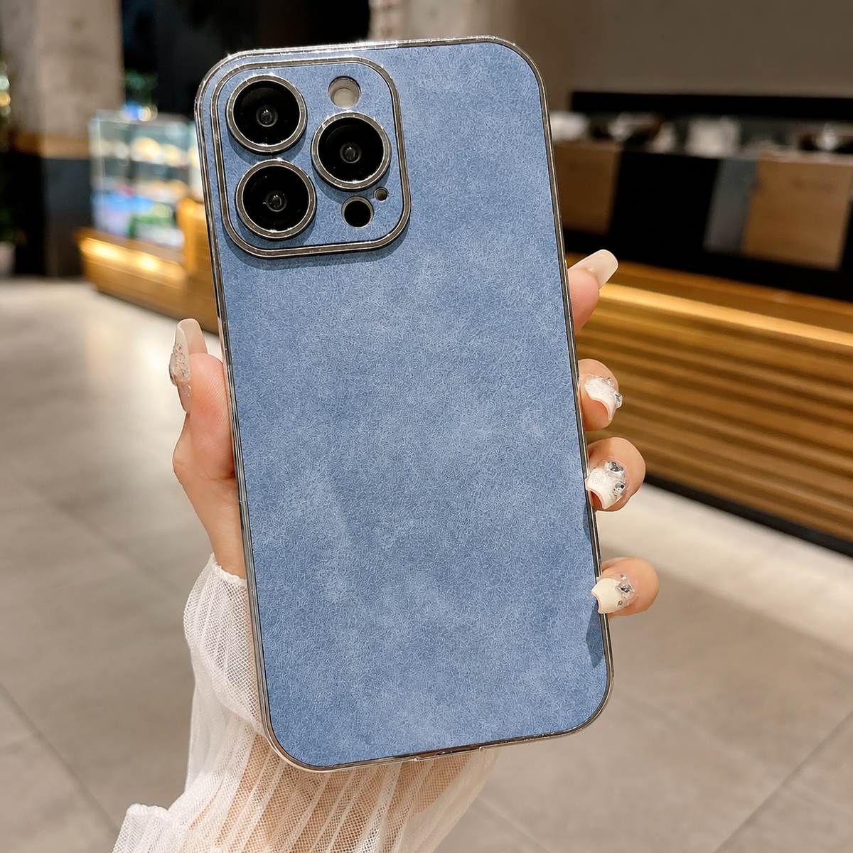 Hand holds a pale blue suede-look case upright in a café. A silver-colored border frames its mottled back and matching camera panel.