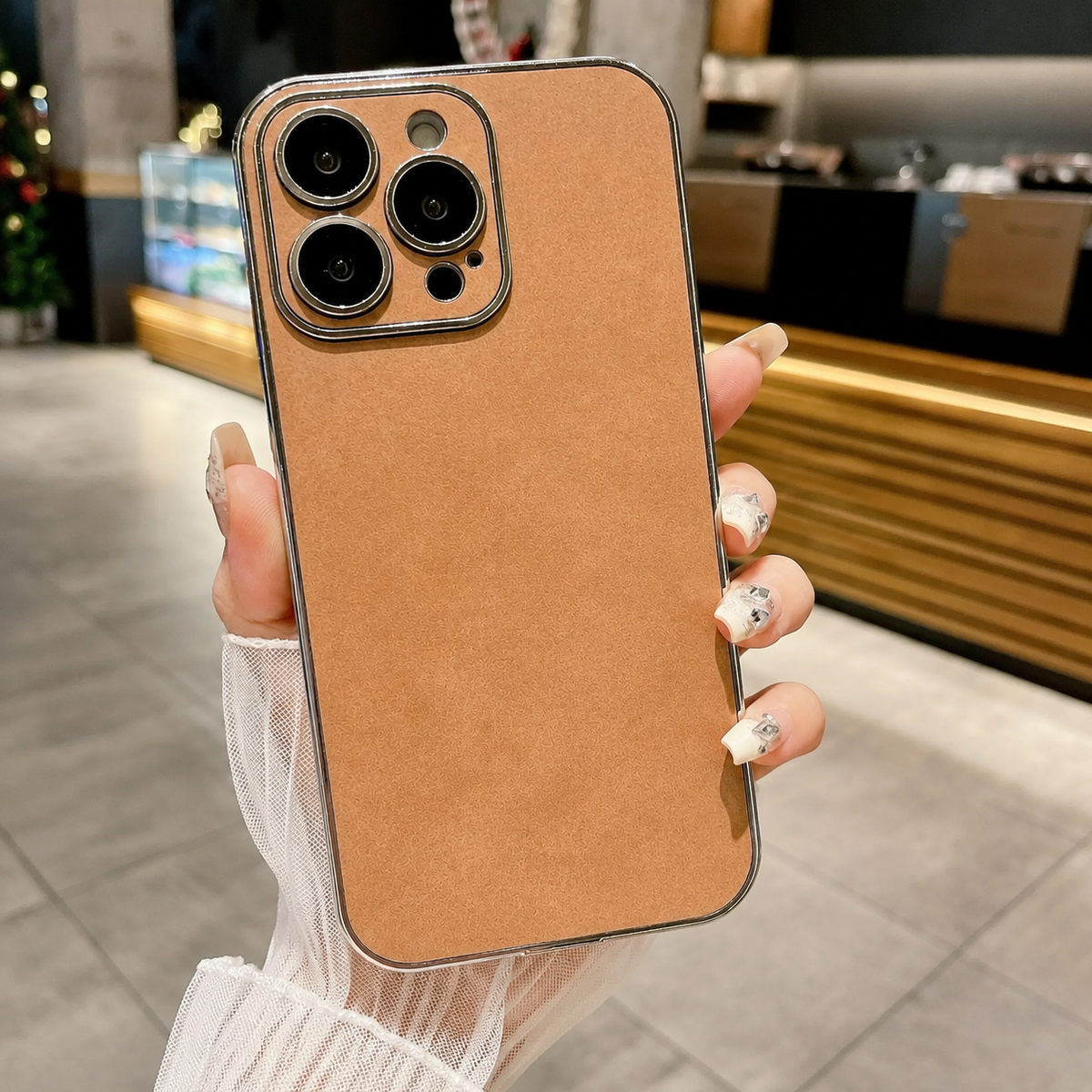 Hand holds a tan suede-look case upright in a café, with a plain mottled back and matching camera panel framed by a silver-colored edge.