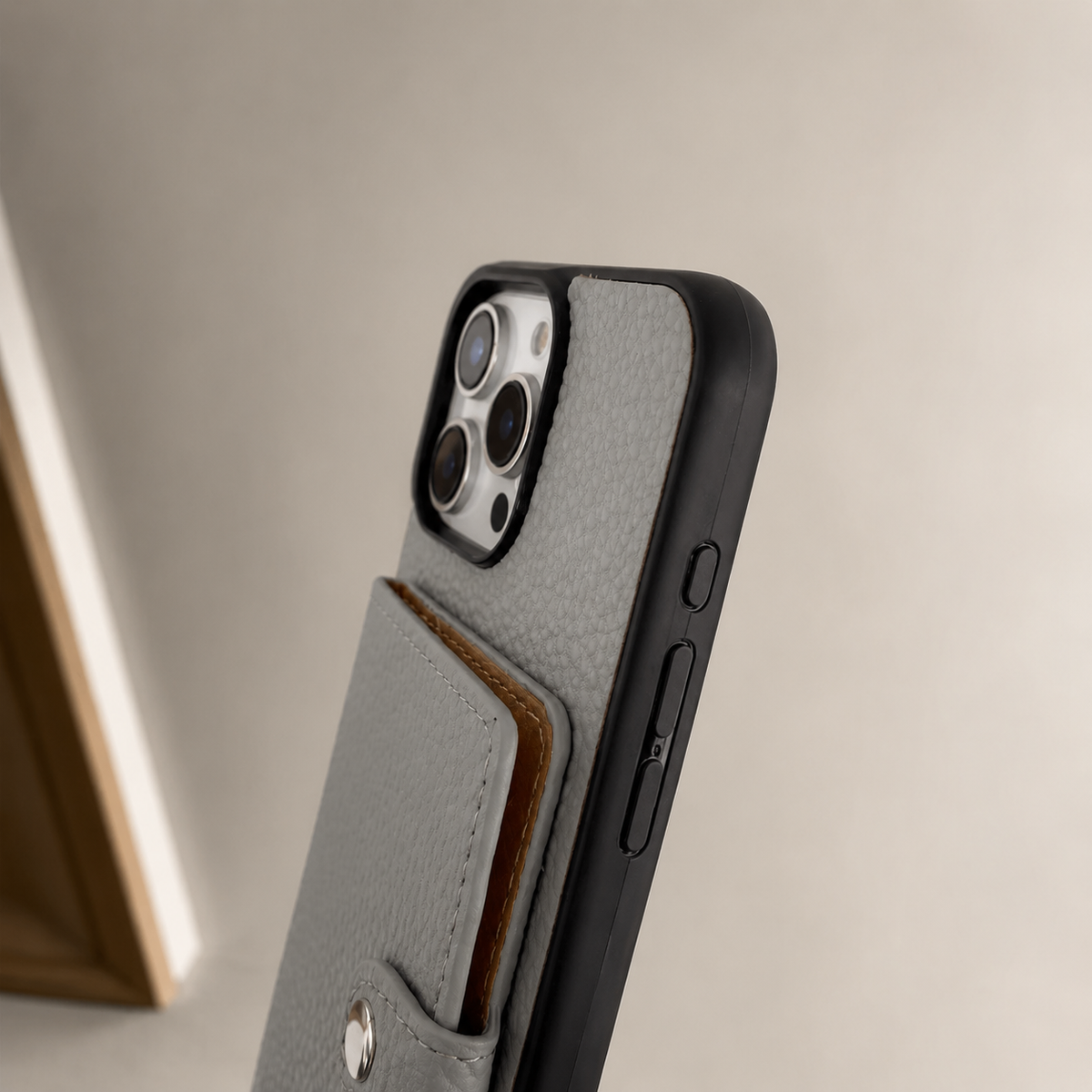 A close side view shows a gray textured wallet phone case standing at an angle against a beige background. The rear card flap projects slightly from the back, with brown card edges visible between the layers. A covered side button and black bumper sit beneath the pale rear camera panel.