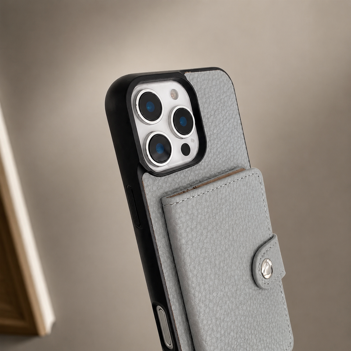A close-up shows the upper rear of a gray textured wallet phone case against a beige background. Three camera lenses sit inside a pale rectangular panel framed by a black bumper. Below, the stitched rectangular card flap is closed with a small silver-colored snap on its tab.