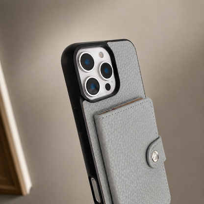 A close-up shows the upper rear of a gray textured wallet phone case against a beige background. Three camera lenses sit inside a pale rectangular panel framed by a black bumper. Below, the stitched rectangular card flap is closed with a small silver-colored snap on its tab.