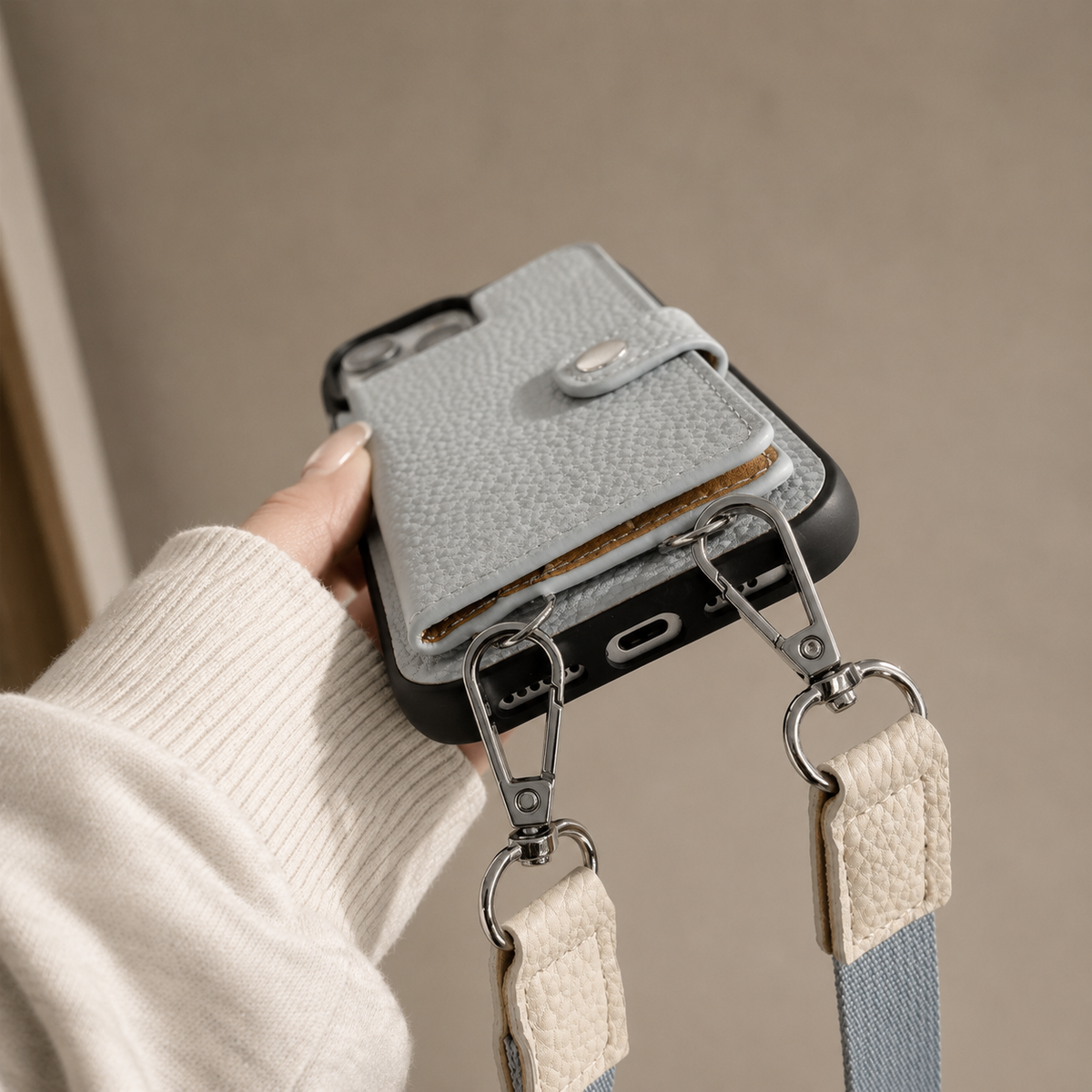 A hand in a pale knit sleeve holds a gray textured wallet phone case with its lower edge tilted toward the viewer. Two black loops are connected to silver-colored strap clips, with pale fabric tabs and blue strap sections hanging below. The rear card flap and snap remain visible.