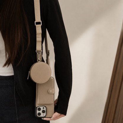 A cropped woman in a black top and dark jeans wears a pale strap carrying a taupe wallet phone case and matching round pouch. The pouch hangs above the phone, whose closed rear card flap faces forward and camera panel sits at the bottom. Her hand rests beside the phone against a beige indoor background.
