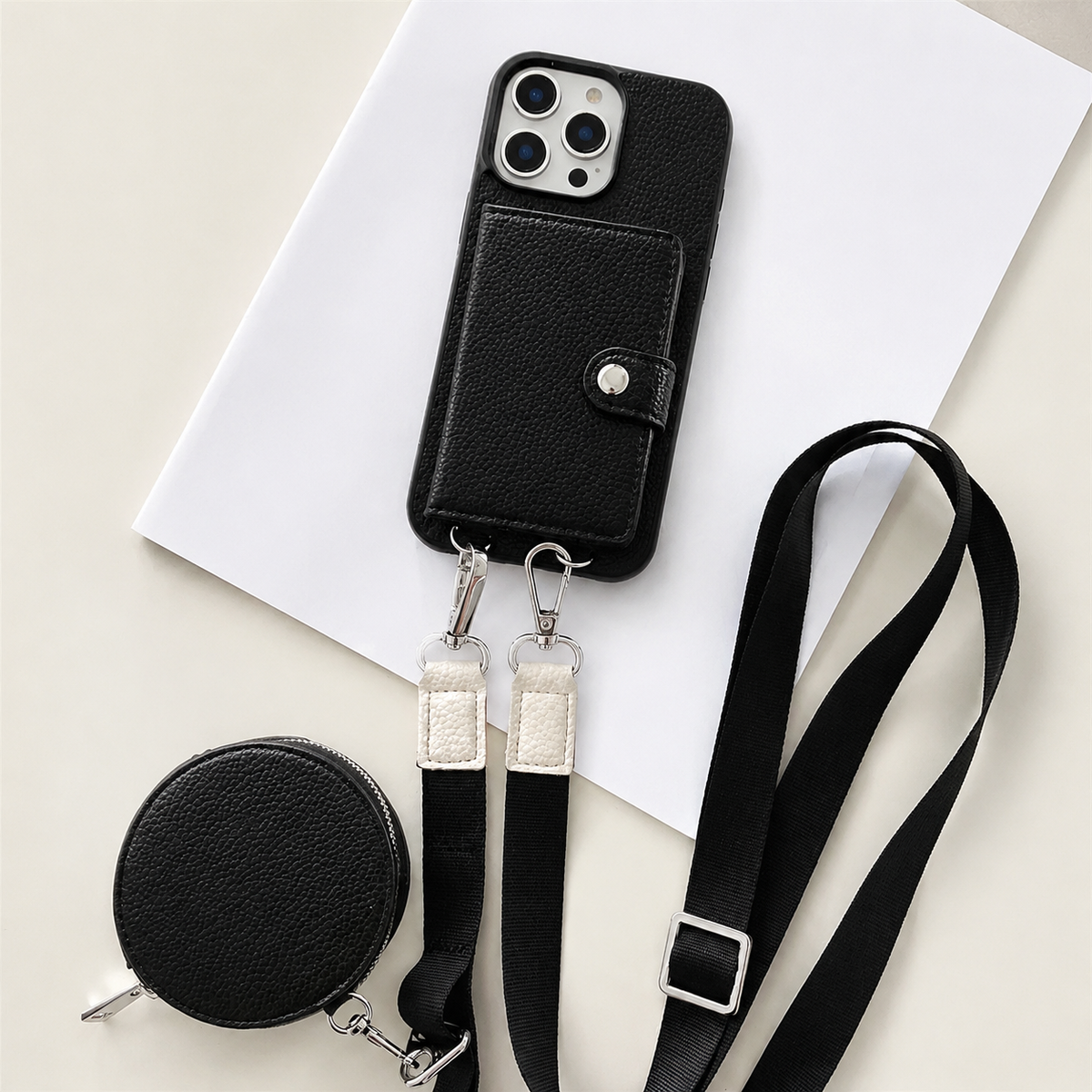 A black textured wallet phone case rests on an angled white sheet beside a matching round zippered pouch and long adjustable strap. A stitched card flap with a silver-colored snap covers the rear. Metal clips connect the strap to two loops on the phone's lower edge, with pale fabric sections near the clips.