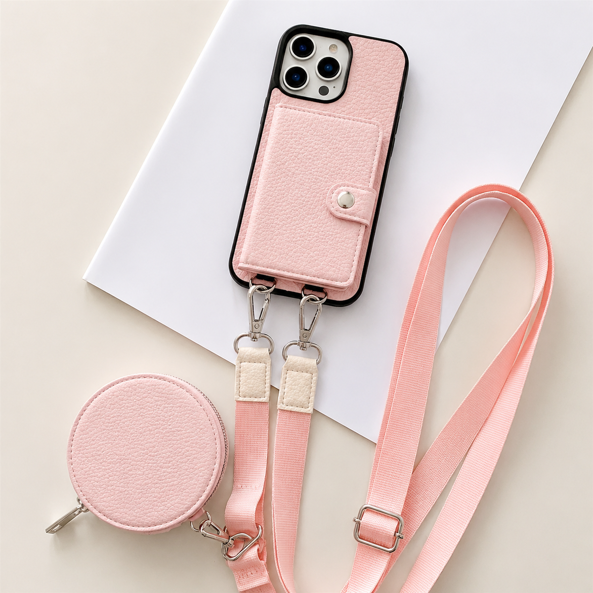 A pale pink textured wallet phone case rests on an angled white sheet beside a matching round zippered pouch and long adjustable strap. A stitched card flap with a silver-colored snap covers the rear. Metal clips connect the strap to two loops on the phone's lower edge, with pale fabric sections near the clips.