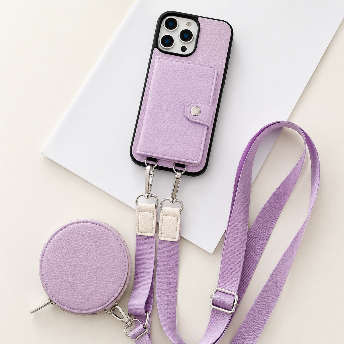 A lavender textured wallet phone case rests on an angled white sheet beside a matching round zippered pouch and long adjustable strap. A stitched card flap with a silver-colored snap covers the rear. Metal clips connect the strap to two loops on the phone's lower edge, with pale fabric sections near the clips.