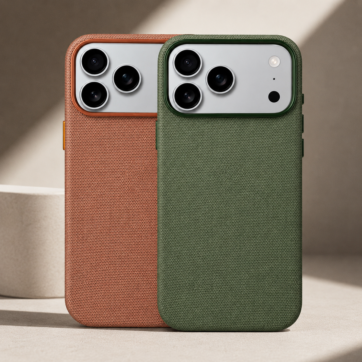 An olive green woven phone case stands in front of a rust brown version on a pale stone surface. Both have silver rectangular camera panels with three black lenses, and matching raised borders around their fabric-covered backs.