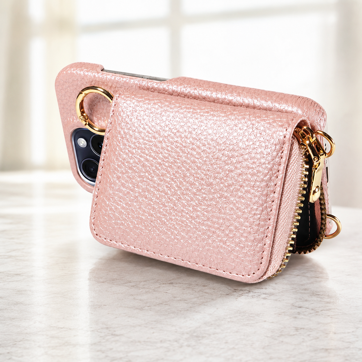 A pale pink pebbled wallet phone case rests sideways on a light countertop. Its rectangular back pouch, gold-colored zipper and ring fitting are visible beside the camera opening.
