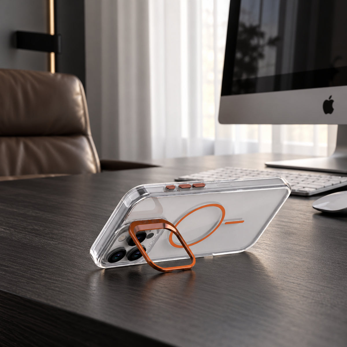 A clear smartphone case with an orange magnetic ring stand, shown resting on a dark wooden desk in front of a computer monitor and keyboard.