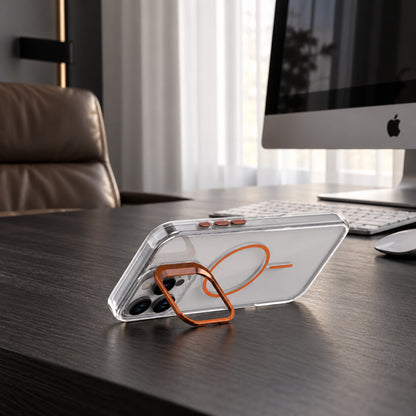 A clear smartphone case with an orange magnetic ring stand, shown resting on a dark wooden desk in front of a computer monitor and keyboard.